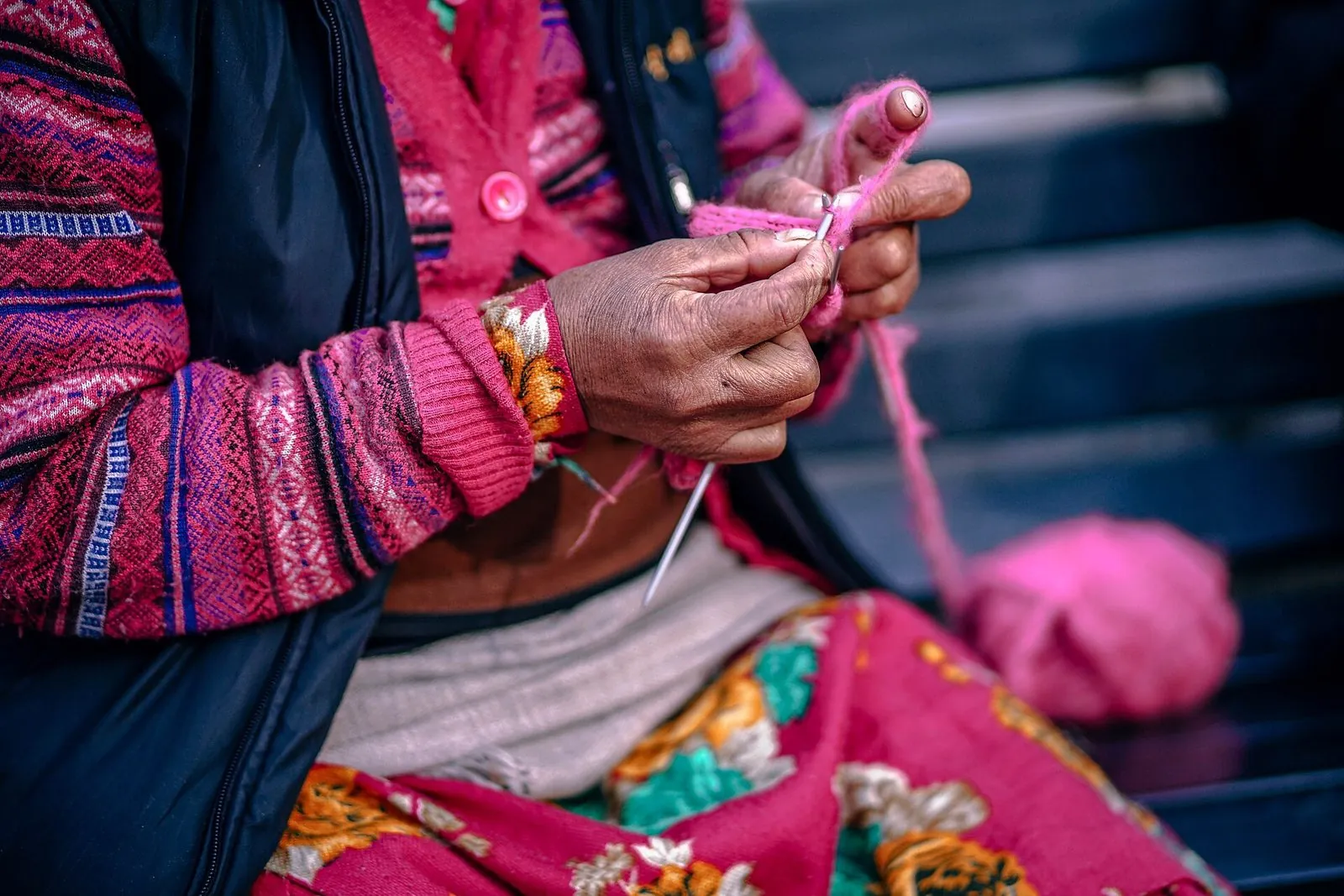 The hands of an older woman in colourful clothes, knitting pink yarn on needles