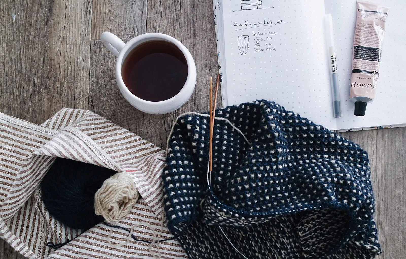 An unfinished navy-and-white speckled knit on needles on a wooden table, beside a cup of tea and an open notebook