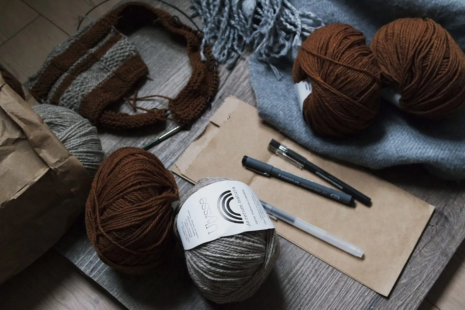 Brown and grey balls of yarn, unfinished knits and pens on a table